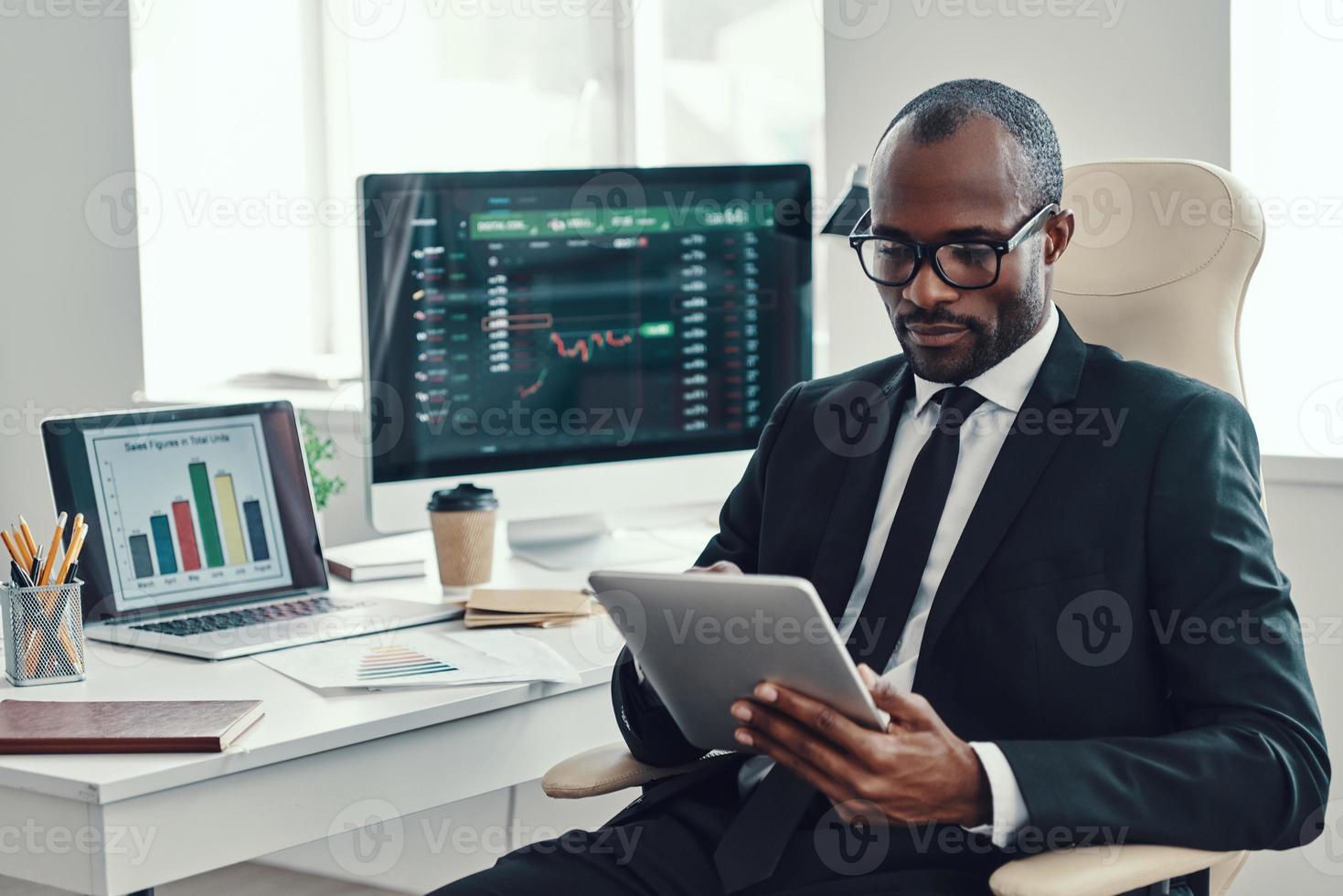 concentrato giovane africano uomo nel vestito formale utilizzando moderno tecnologie mentre Lavorando nel il ufficio foto