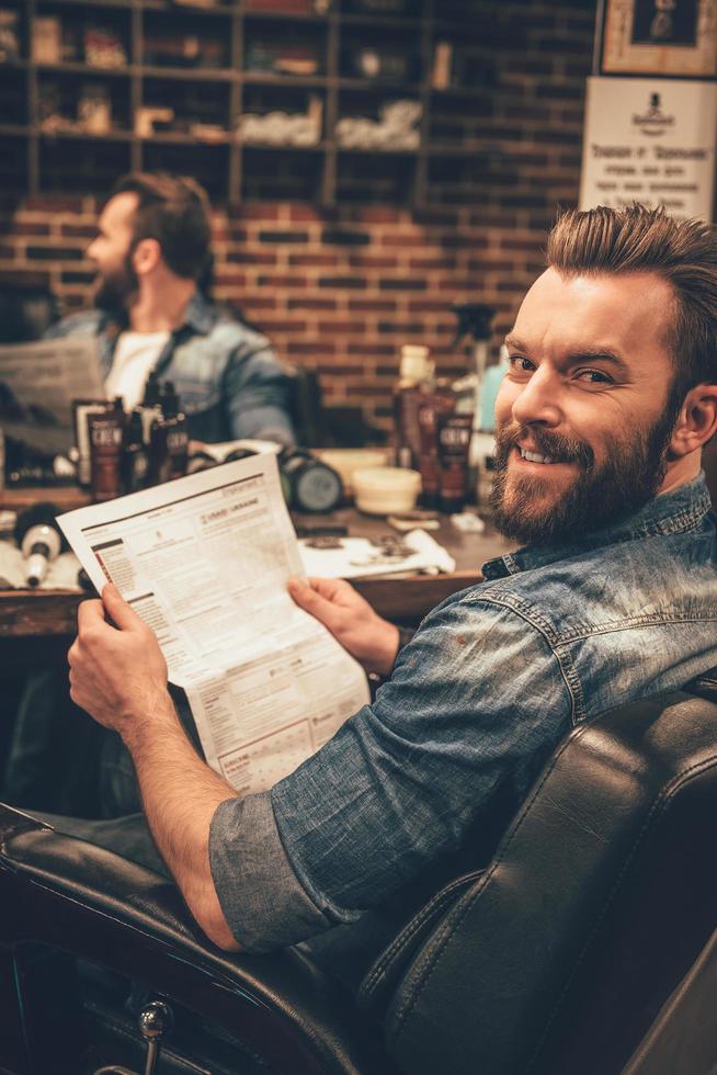 pronto per nuovo taglio di capelli. bello giovane barbuto uomo al di sopra di spalla con Sorridi e Tenere giornale mentre seduta nel sedia a barbiere foto