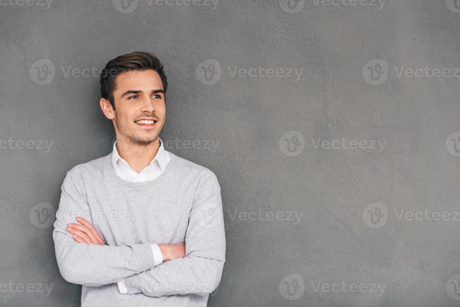 guardare nel futuro con Sorridi. fiducioso giovane uomo conservazione braccia attraversato e guardare lontano con Sorridi mentre in piedi contro grigio sfondo foto