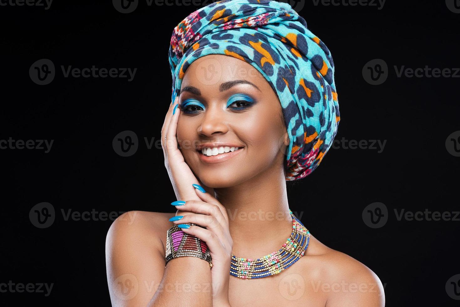 nel amore con sua stile. bellissimo africano donna indossare un' foulard e gioielleria e sorridente mentre in piedi contro nero sfondo foto