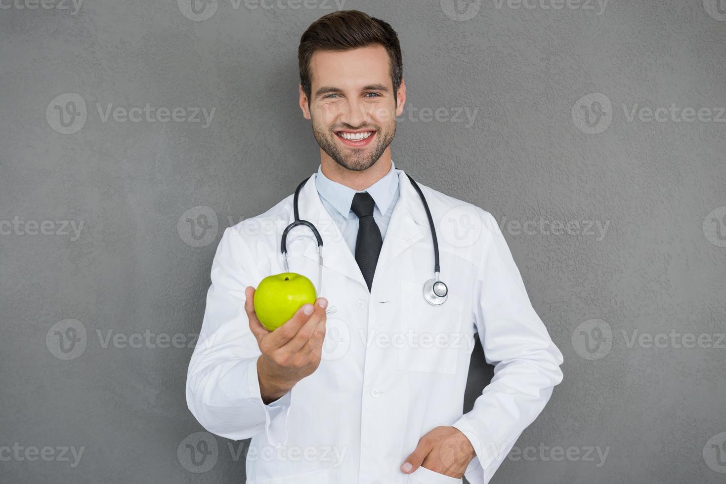 vitamine siamo importante per Salute. allegro giovane medico nel bianca uniforme allungamento su verde Mela e sorridente mentre in piedi contro grigio sfondo foto