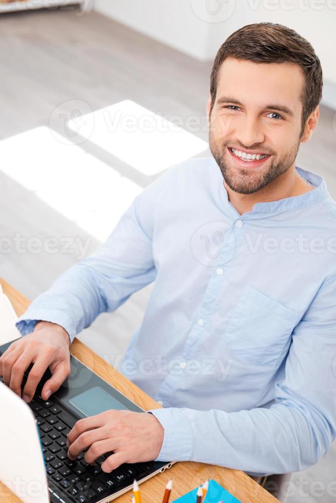 assunzione cura di attività commerciale con Sorridi. superiore Visualizza di bello giovane uomo nel camicia Lavorando su il computer portatile e sorridente a telecamera mentre seduta a il suo Lavorando posto foto