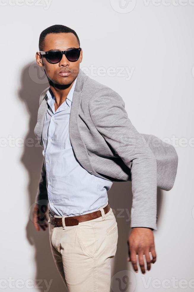 freddo e di tendenza. fiducioso giovane africano uomo nel occhiali da sole indossare giacca e guardare al di sopra di spalla mentre in piedi contro bianca sfondo foto