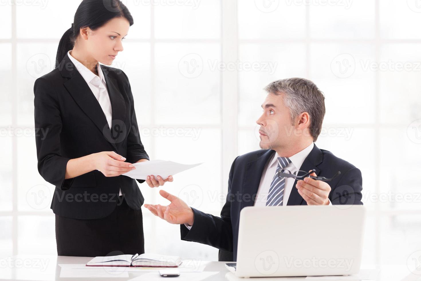 Lavorando su i problemi. frustrato maturo uomo d'affari seduta a il tavolo e Esprimere a gesti mentre giovane donna nel vestito formale Tenere documenti foto