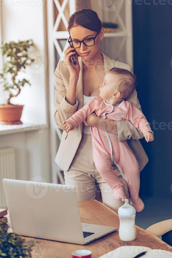 Lavorando madre con bambino. giovane bellissimo donna d'affari parlando su mobile Telefono e guardare a il computer portatile mentre in piedi con sua bambino ragazza a sua Lavorando posto foto