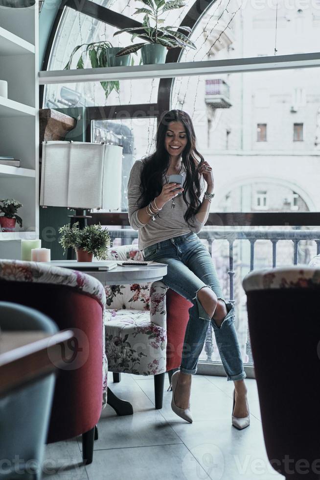 chiacchierando con gli amici. pieno lunghezza di bellissimo giovane sorridente donna utilizzando sua inteligente Telefono mentre pendente su il sedia nel ristorante foto