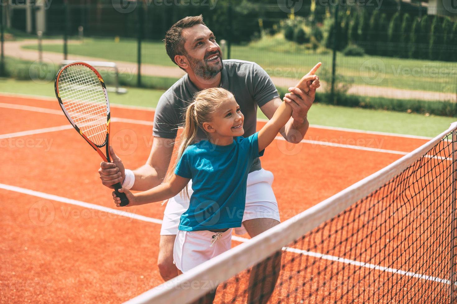 tennis è divertimento quando padre è vicino. allegro padre nel gli sport capi di abbigliamento insegnamento il suo figlia per giocare tennis mentre tutti e due in piedi su tennis Tribunale foto