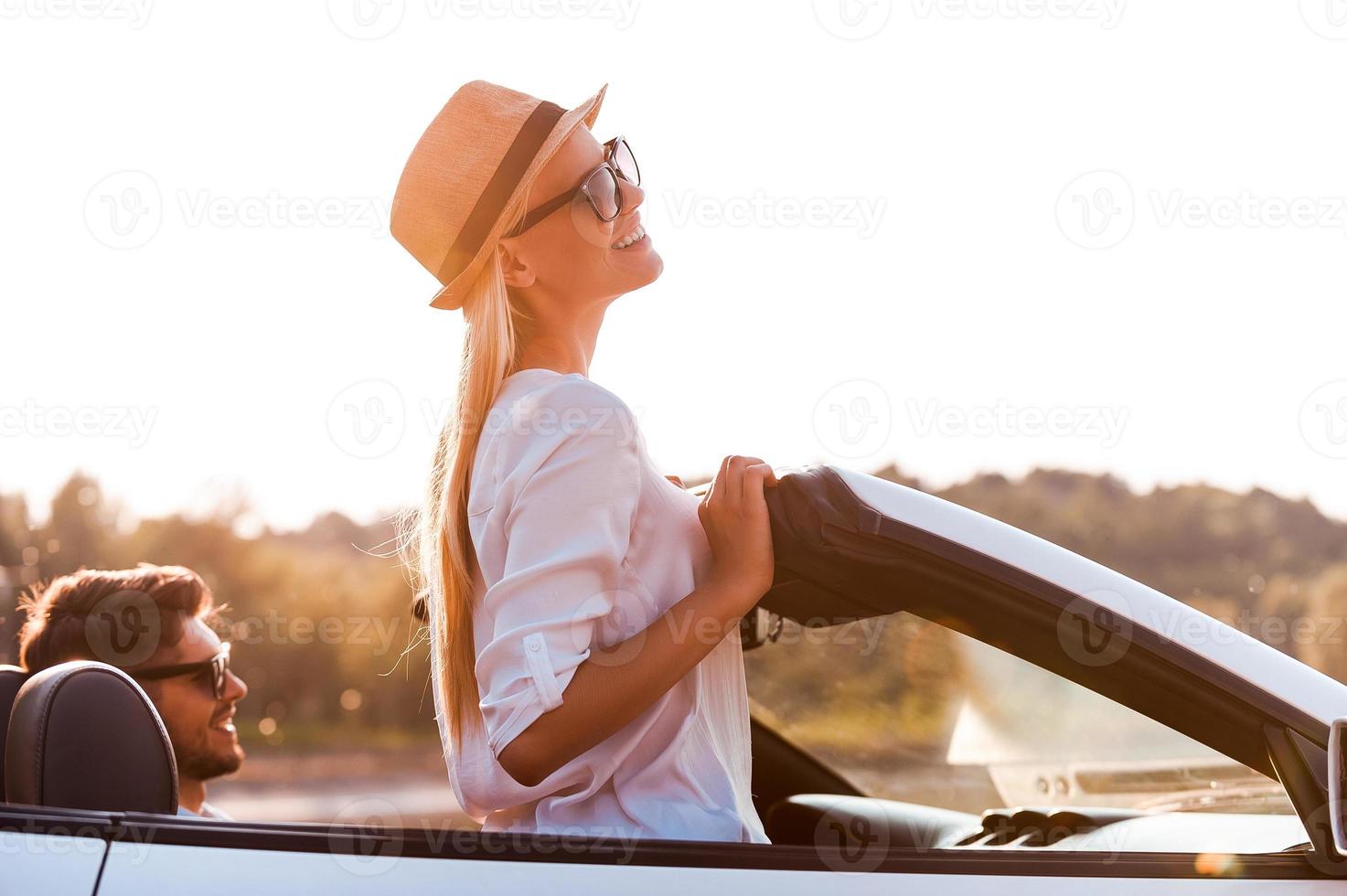 godendo il sole e fresco aria. lato Visualizza di allegro giovane donna pendente a il vento scudo mentre sua fidanzato seduta vicino su davanti posto a sedere di convertibile foto