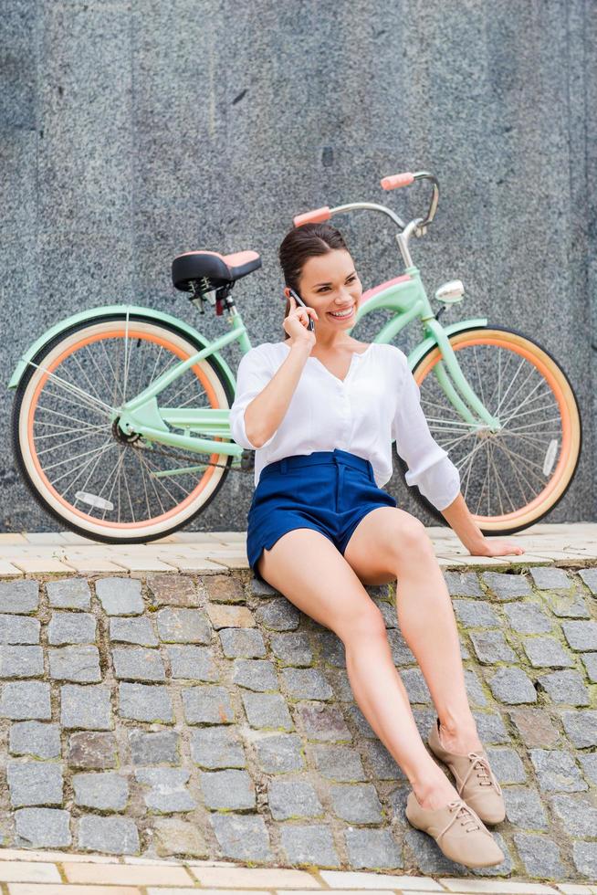 spensierato e elegante. bellissimo giovane sorridente donna parlando su il mobile Telefono mentre seduta a il ciglio della strada vicino sua Vintage ▾ bicicletta foto