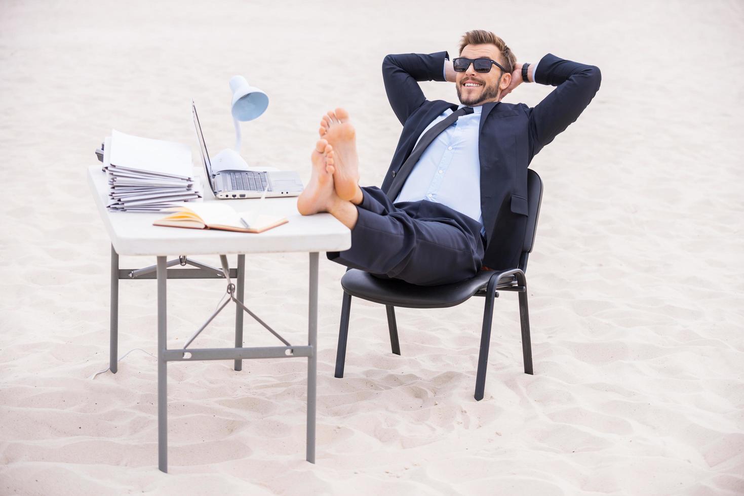 pronto per vacanza. bello giovane uomo nel vestito formale e occhiali da sole Tenere mani dietro a testa e Tenere il suo piedi su il tavolo in piedi su sabbia foto