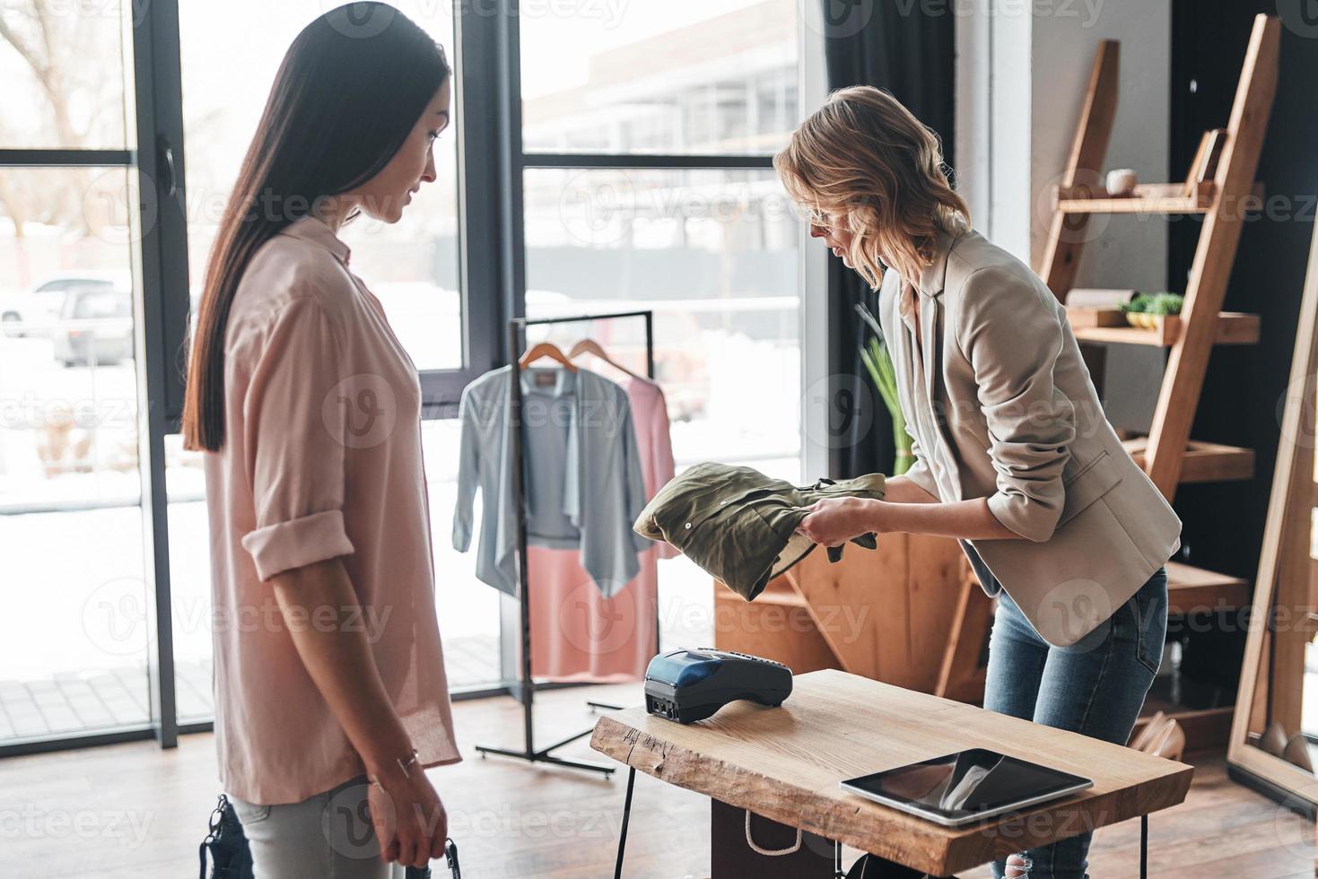 assistere. bellissimo giovane donna pieghevole un' camicia per sua cliente mentre Lavorando nel il moda boutique foto