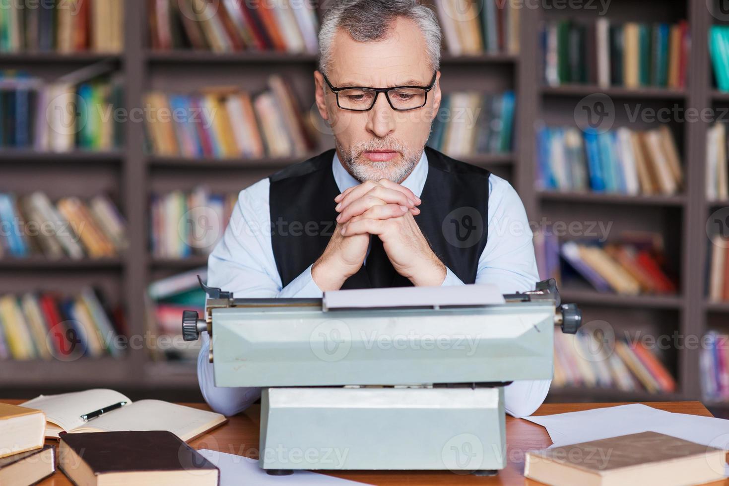 in attesa per ispirazione. fiducioso grigio capelli anziano uomo nel vestito formale seduta a il macchina da scrivere e guardare concentrato con scaffale nel il sfondo foto