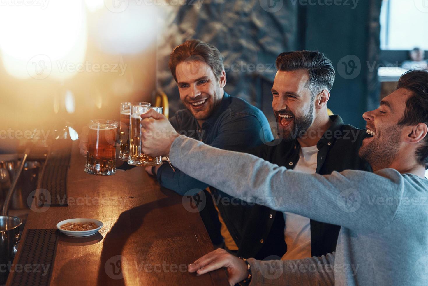 contento giovane uomini nel casuale capi di abbigliamento tostatura ogni altro con birra e ridendo mentre seduta nel il pub foto