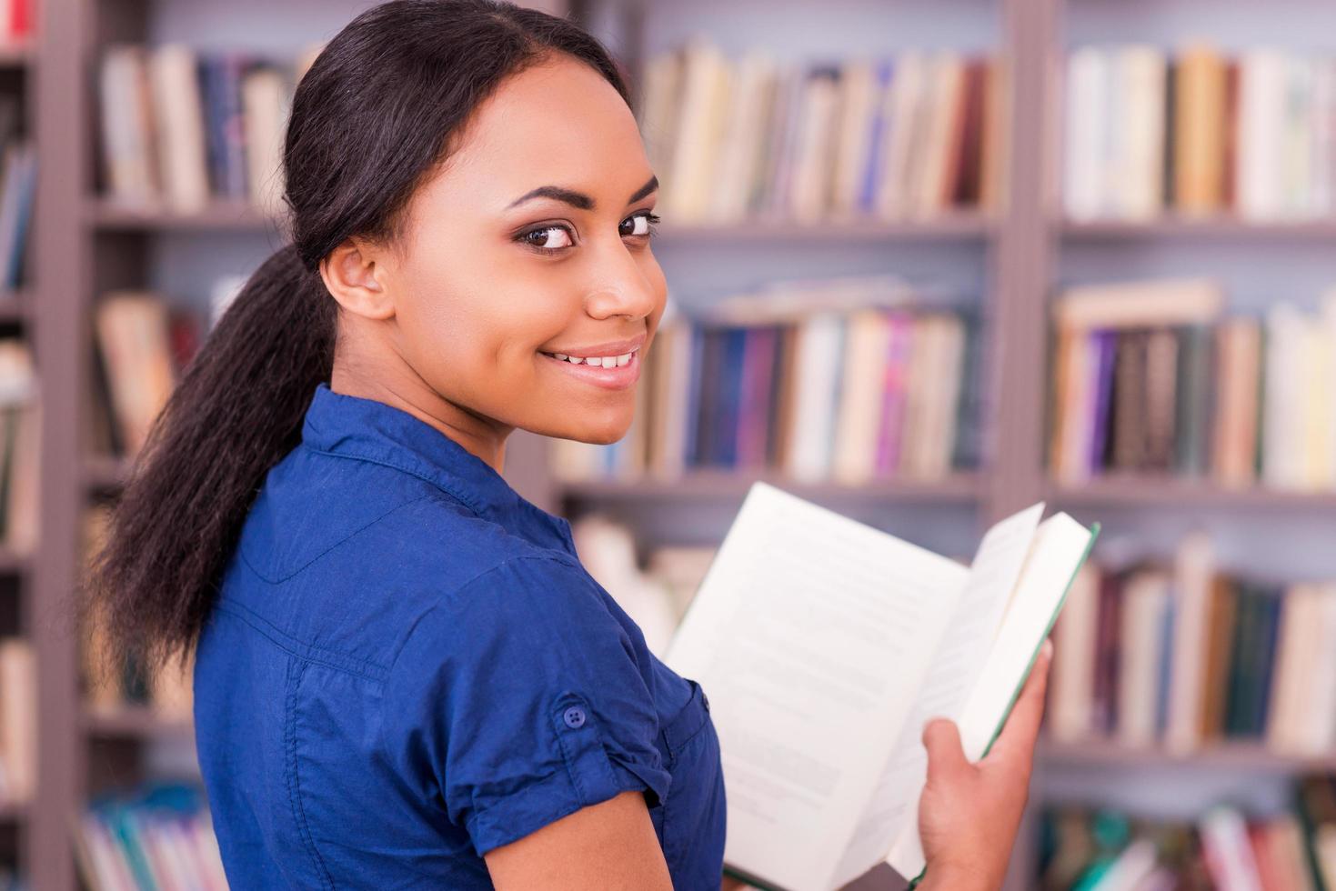 lettura sua preferito libro. posteriore Visualizza di bellissimo africano femmina alunno Tenere un' libro e guardare al di sopra di spalla con Sorridi mentre in piedi nel biblioteca foto