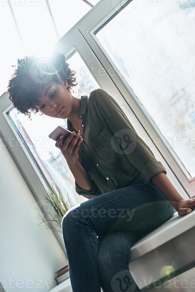 appena chattare. attraente giovane africano donna utilizzando sua inteligente Telefono e sorridente mentre seduta su il finestra davanzale in casa foto