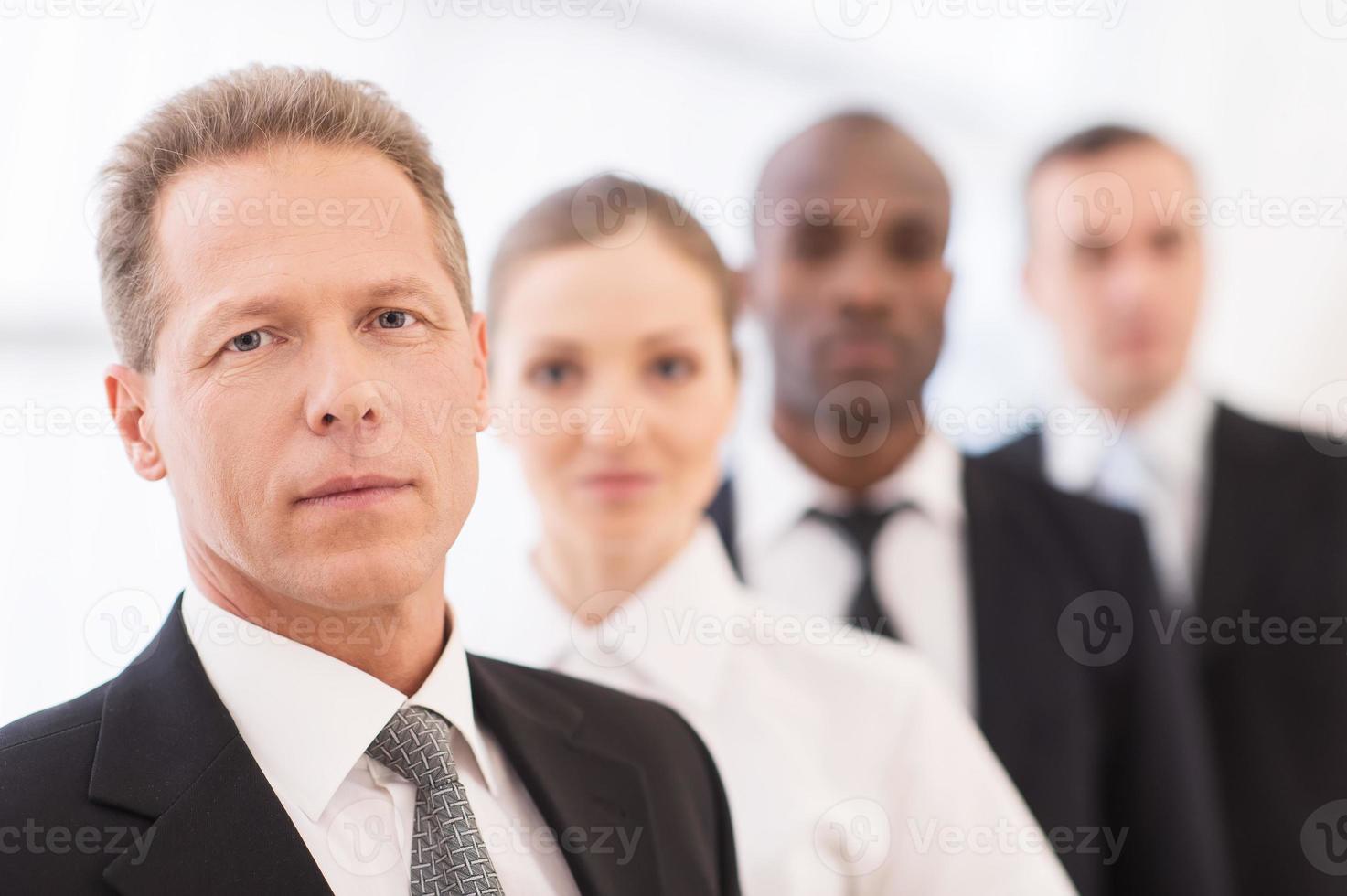 attività commerciale squadra. fiducioso maturo uomo nel vestito formale guardare a telecamera mentre il suo colleghi in piedi nel linea dietro a lui foto