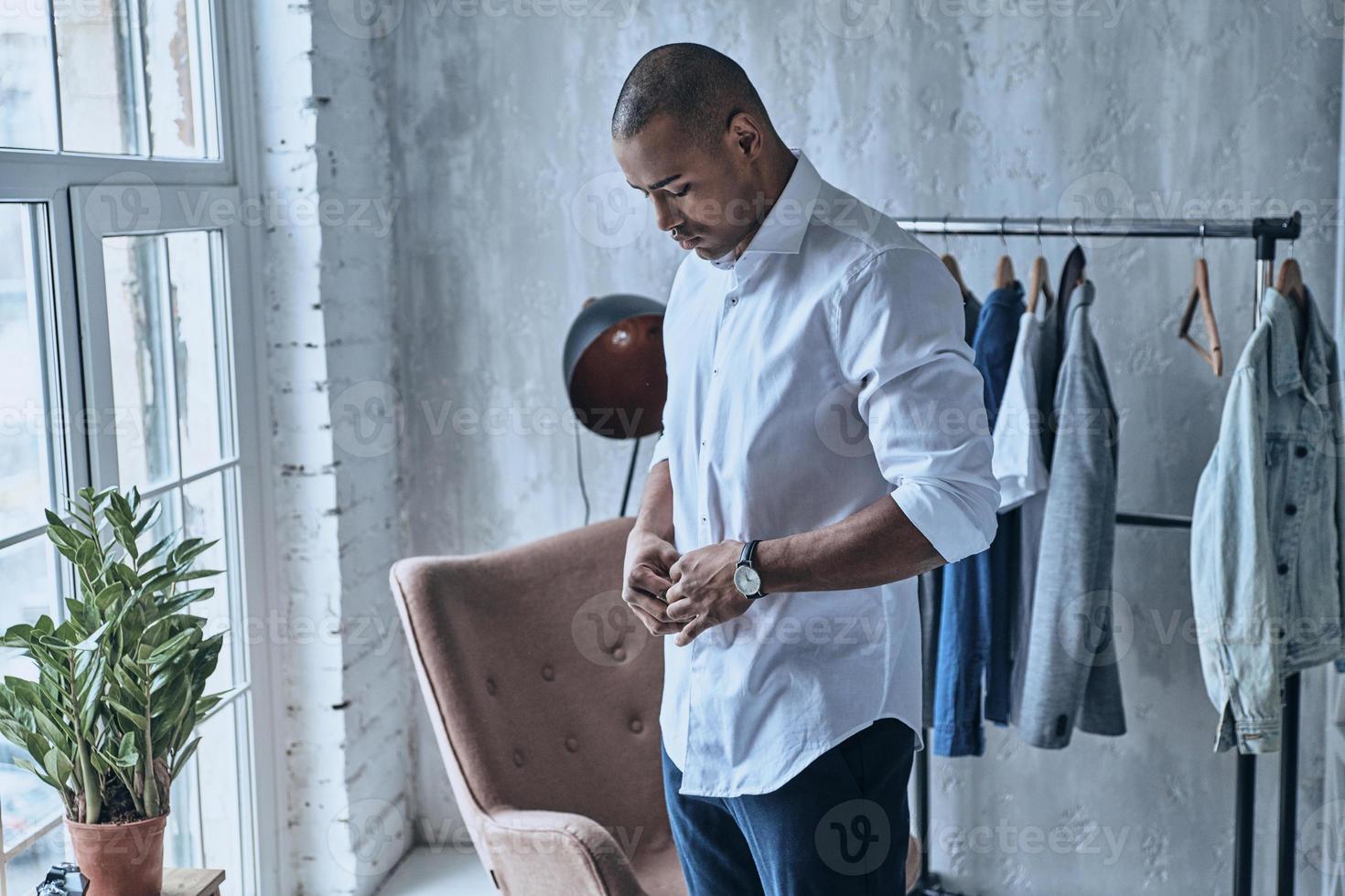 pagare Attenzione per ogni dettaglio. bello giovane africano uomo abbottonatura il suo camicia mentre in piedi a casa foto