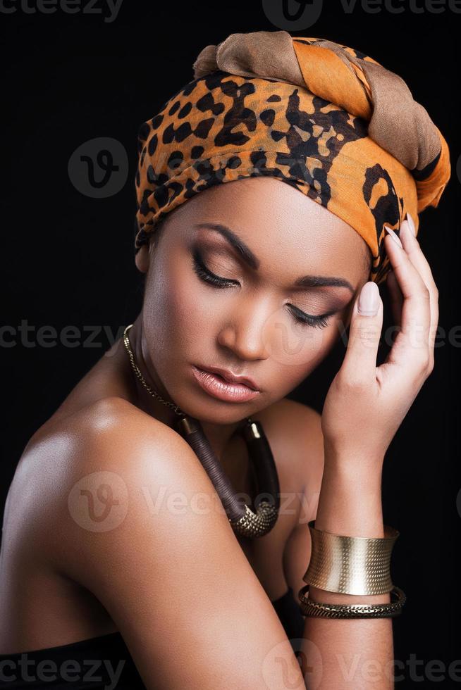 bellezza su nero. bellissimo africano donna indossare un' foulard e toccante testa mentre in piedi contro nero sfondo foto