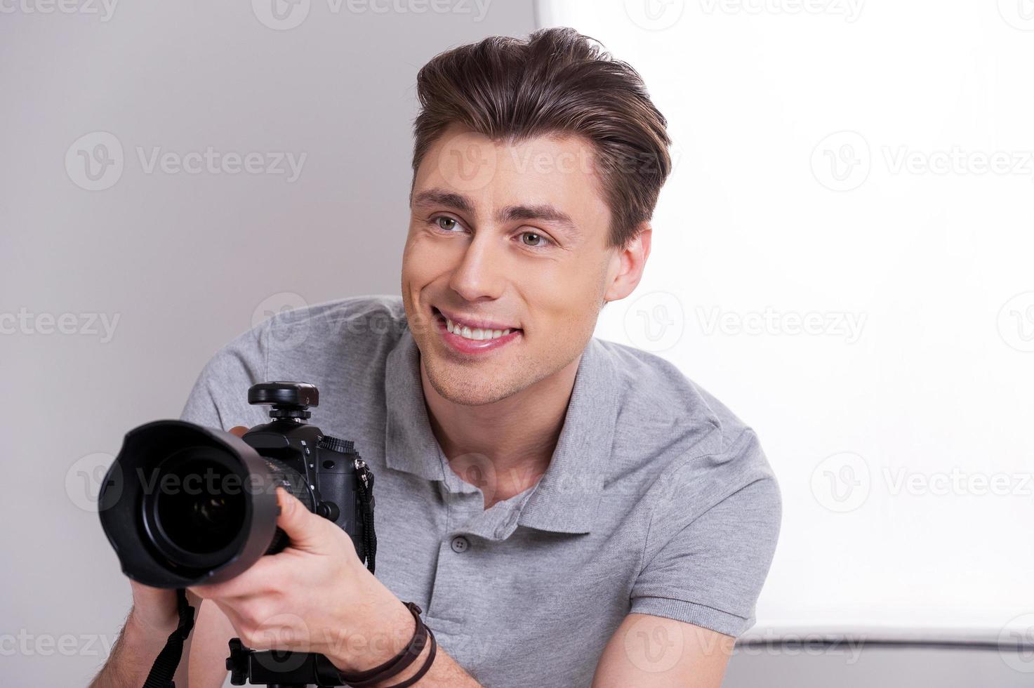 fotografo a opera. giovane uomo nel polo camicia Tenere digitale telecamera e guardare lontano mentre in piedi nel studio con illuminazione attrezzatura su sfondo fotografo a opera. giovane uomo nel polo camicia Tenere digitale telecamera e guardare lontano mentre in piedi nel st foto