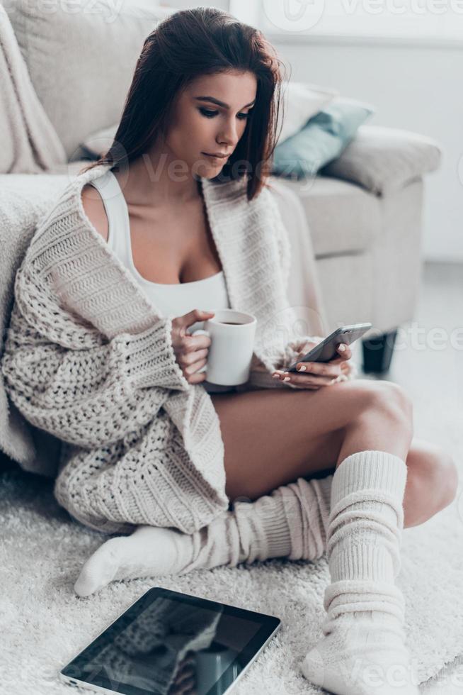 gratuito tempo a casa. bellissima giovane donna guardare a mobile Telefono e Tenere un' tazza mentre seduta su tappeto a casa foto
