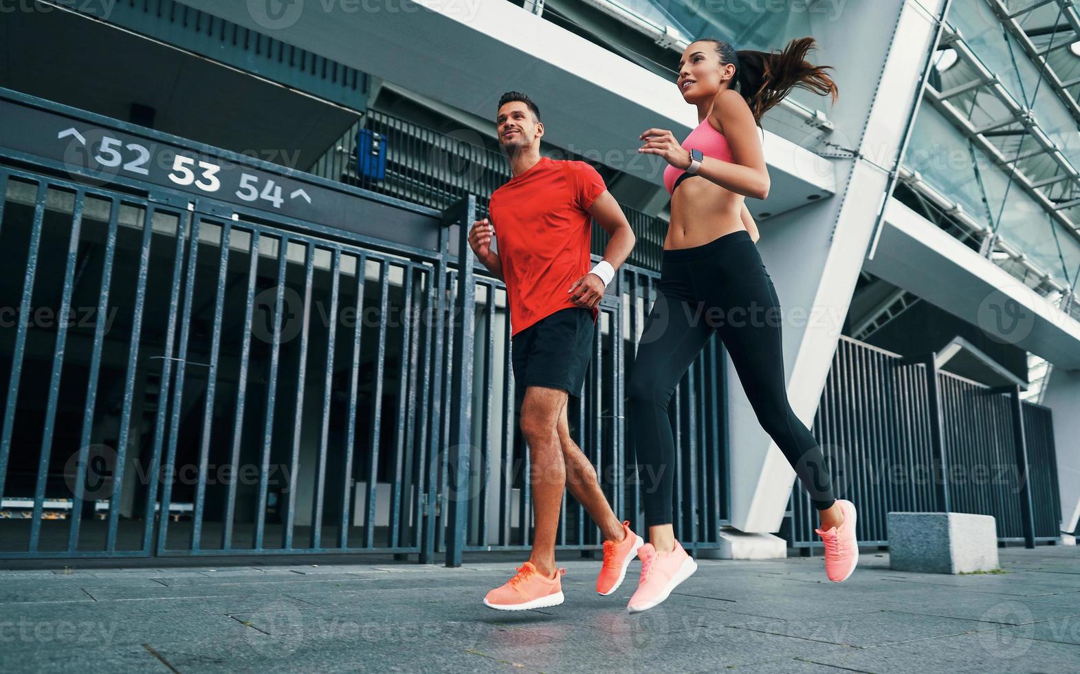 pieno lunghezza di fiducioso giovane uomo e donna nel gli sport capi di abbigliamento in esecuzione e sorridente mentre praticante all'aperto foto