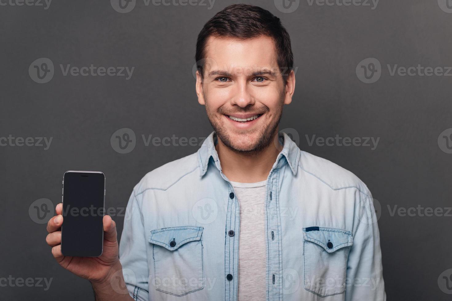 avere voi visto Questo allegro giovane uomo spingendo il suo inteligente Telefono con copia spazio e guardare a telecamera con Sorridi mentre in piedi contro grigio sfondo foto