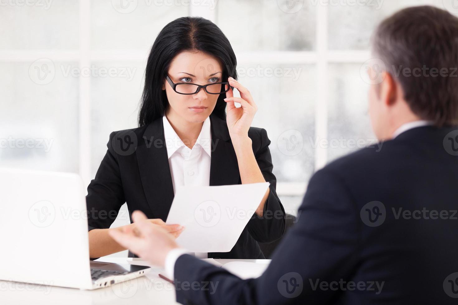 lavoro colloquio. fiducioso giovane donna nel vestito formale Tenere un' carta e regolazione sua bicchieri mentre grigio capelli uomo seduta nel davanti di sua e Esprimere a gesti foto