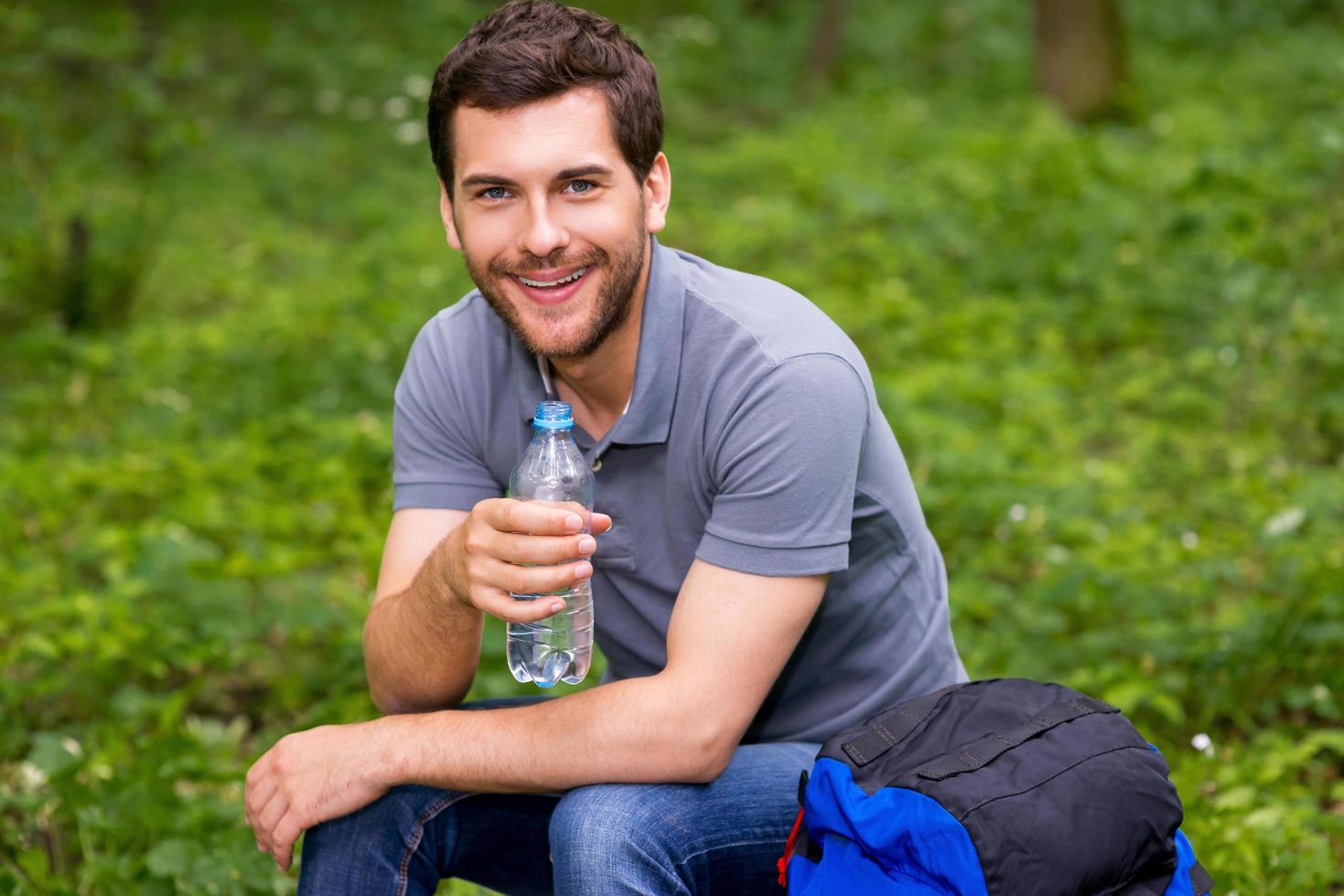 assetato viaggiatore. contento giovane uomo Tenere bottiglia con acqua mentre seduta nel un' foresta con zaino posa vicino lui con foto