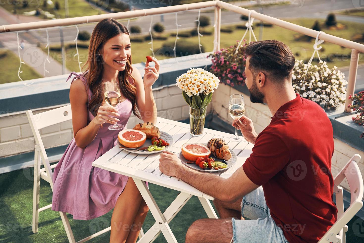 superiore Visualizza di spensierato giovane coppia nel casuale capi di abbigliamento godendo romantico cena e sorridente mentre seduta su il tetto terrazza all'aperto foto