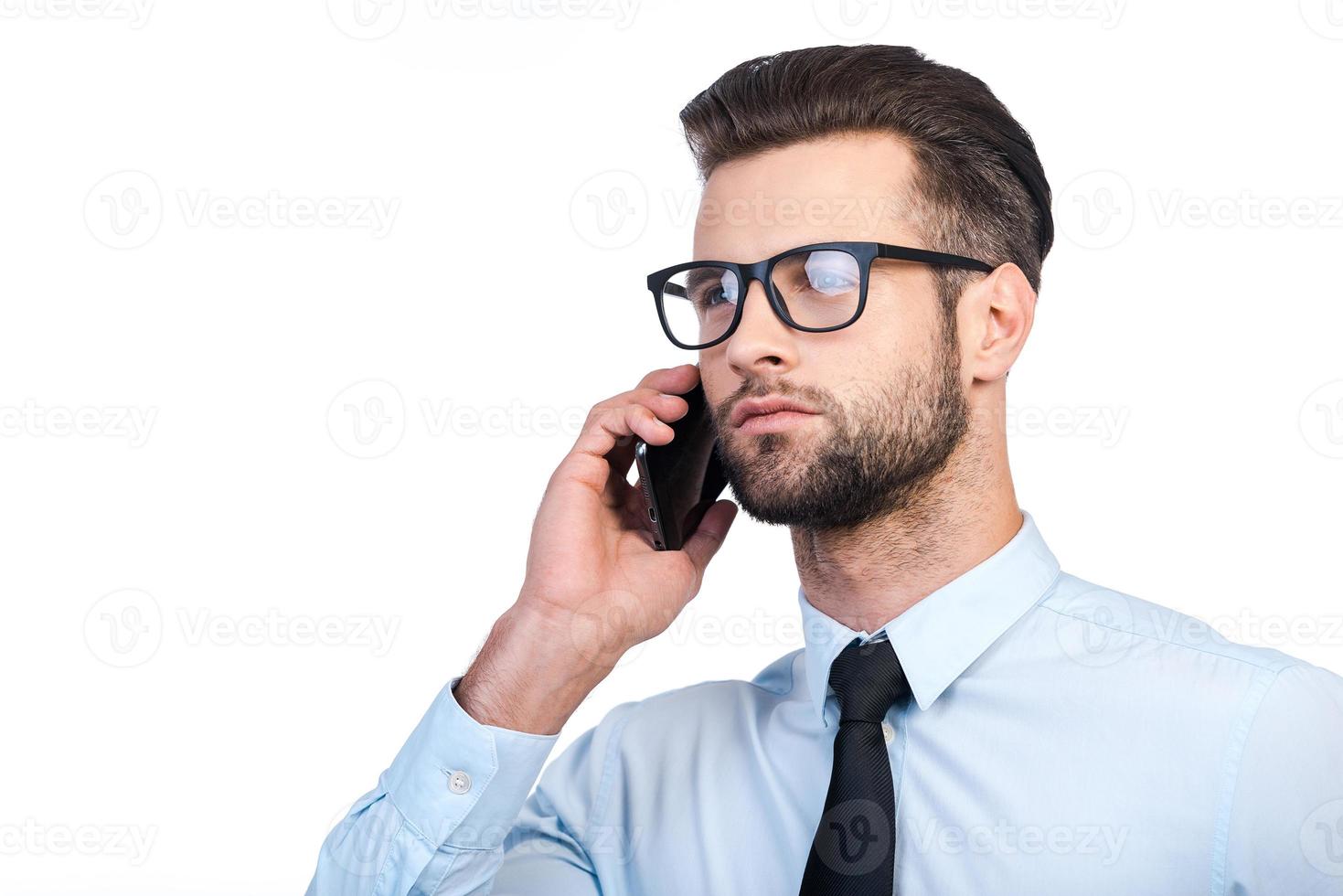 soggiorno nel tocco. fiducioso giovane bello uomo nel camicia e cravatta parlando su mobile Telefono mentre in piedi contro bianca sfondo foto