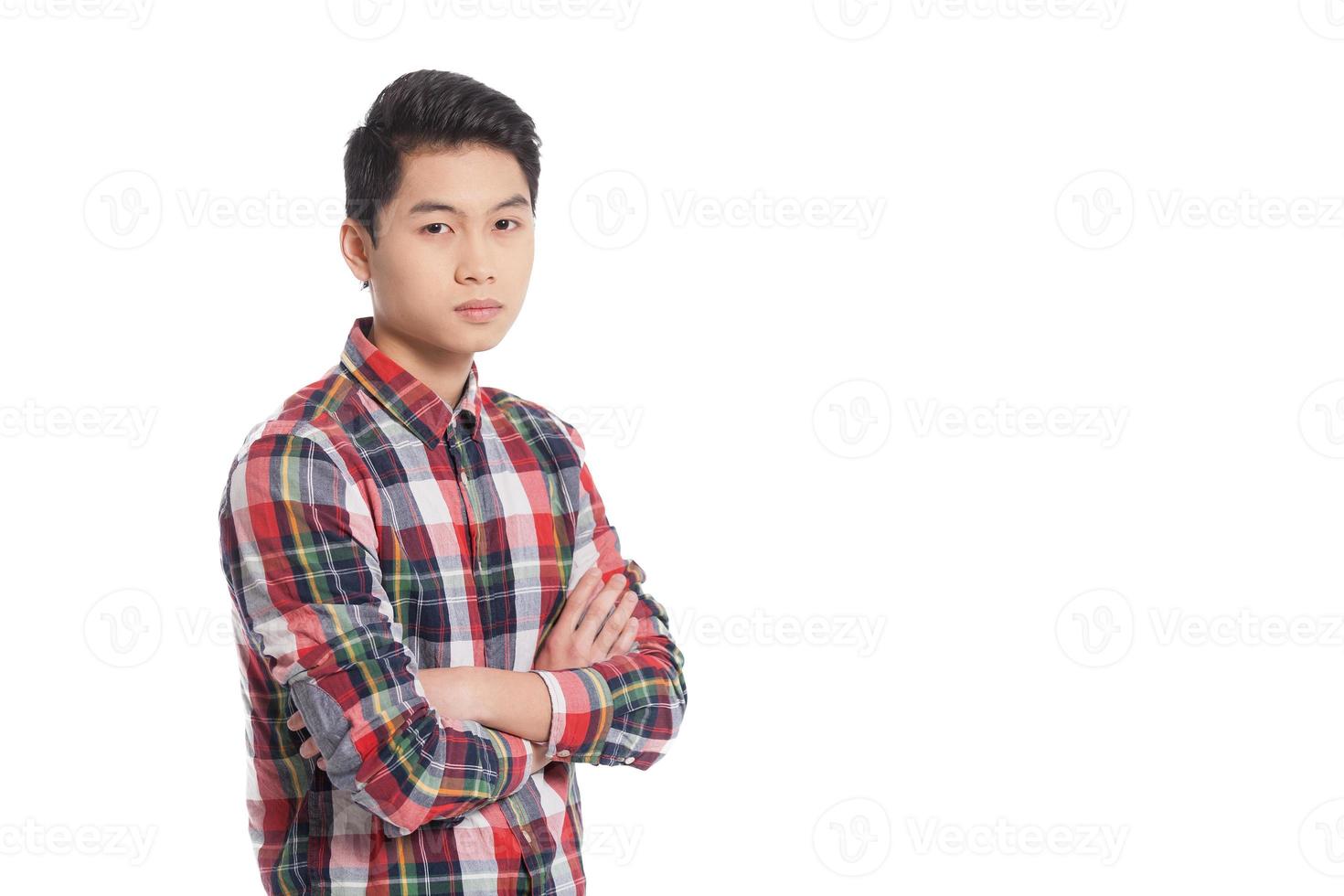fiducioso Guarda. sorridente Cinese adolescenziale ragazzo conservazione braccia attraversato e sorridente mentre in piedi isolato su bianca foto