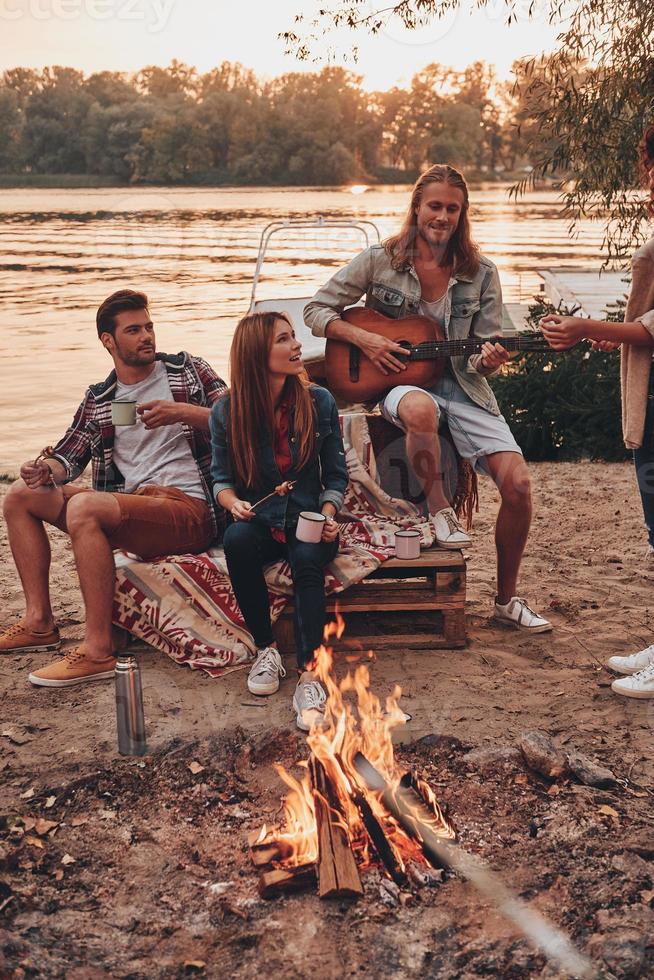 sorprendente sera. gruppo di giovane persone nel casuale indossare sorridente mentre godendo spiaggia festa vicino il fuoco di bivacco foto