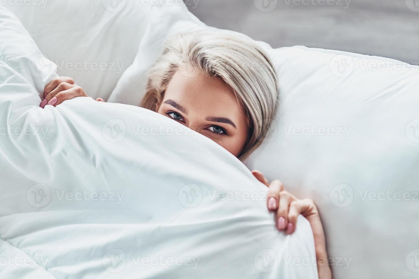 pure pigro per scia su. superiore Visualizza di attraente giovane donna copertura metà di sua viso con coperta e guardare a telecamera mentre dire bugie nel letto a casa foto