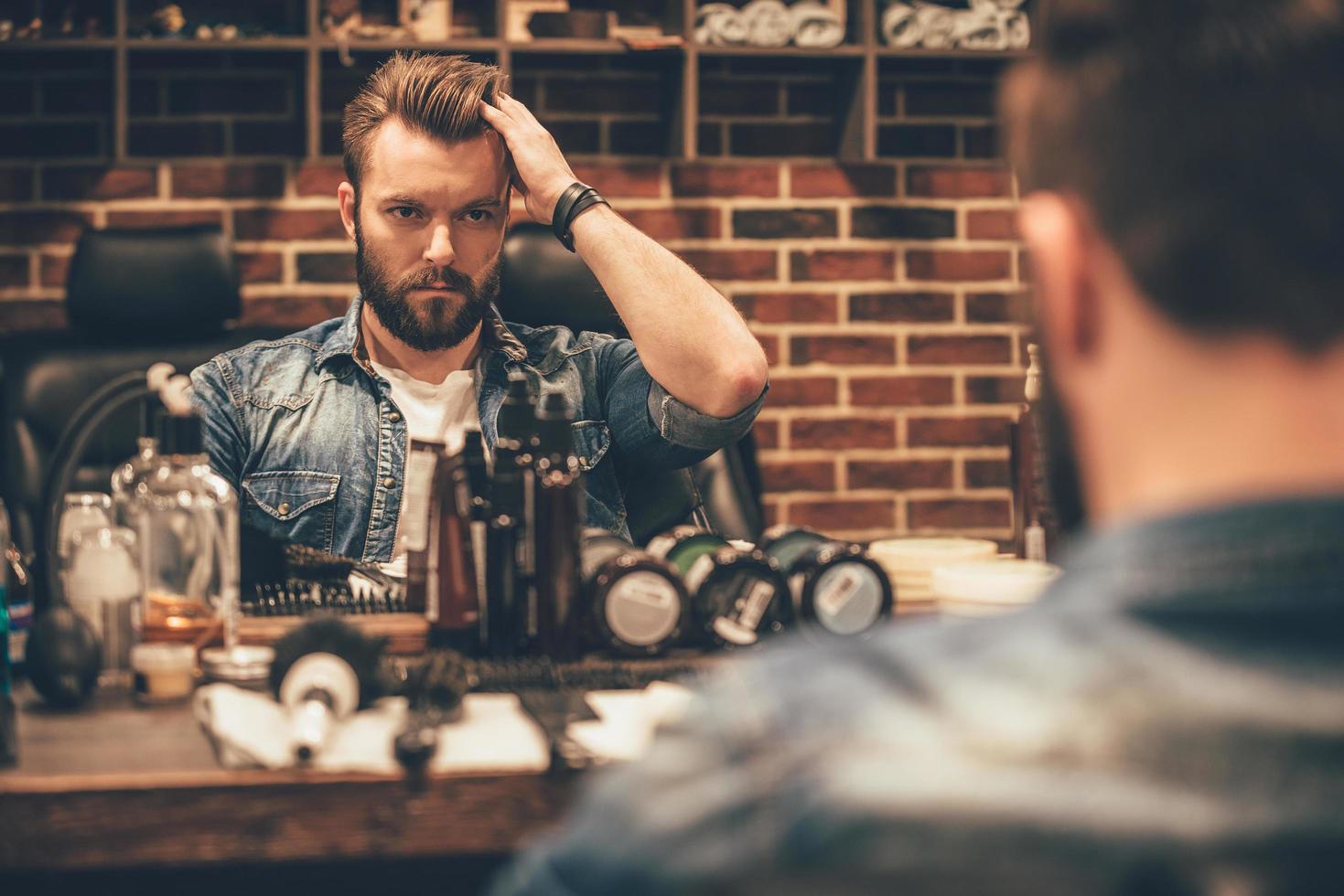 tempo per nuovo taglio di capelli. bello giovane barbuto uomo guardare a il suo riflessione nel il specchio e conservazione mano nel capelli mentre seduta nel sedia a barbiere foto