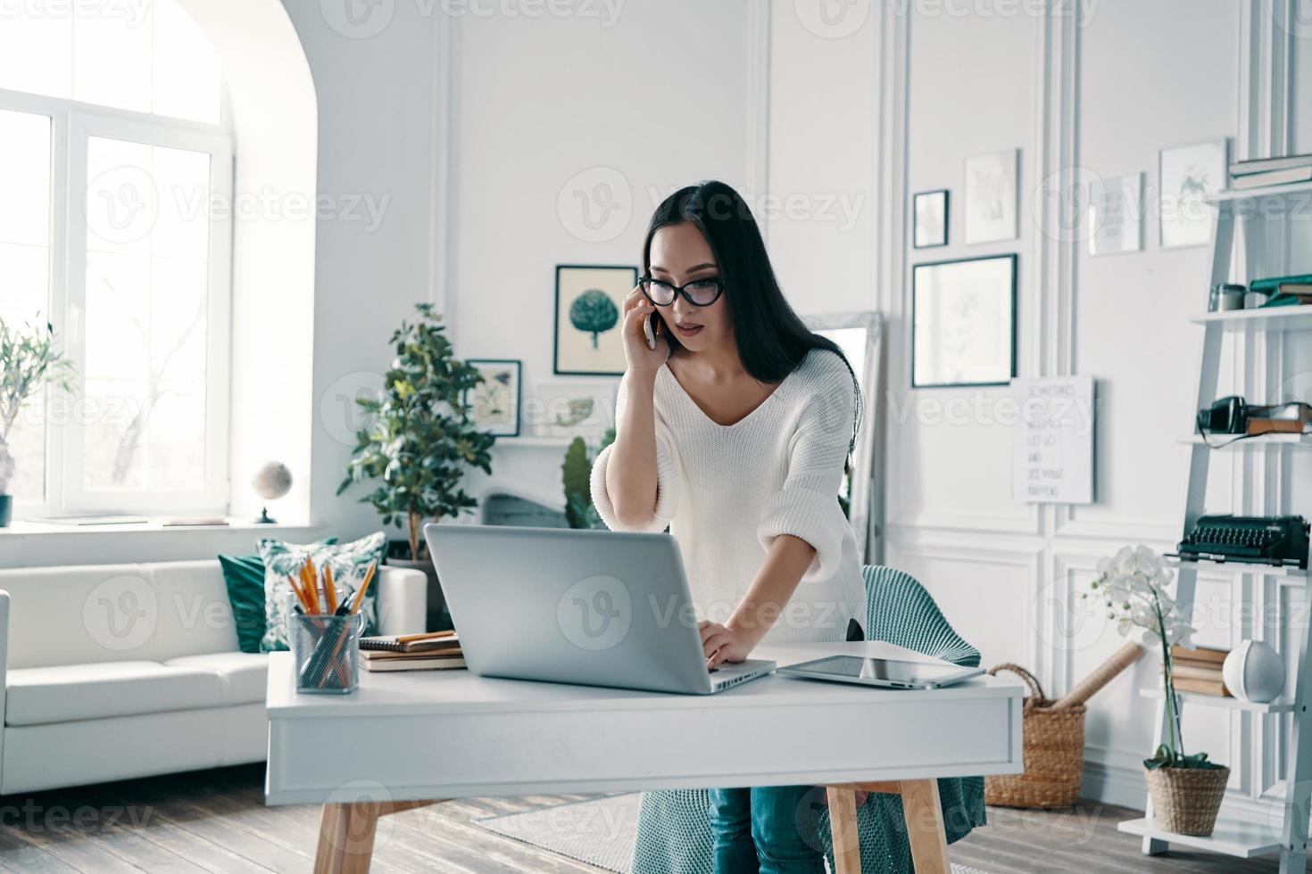 sempre a disposizione. bellissimo giovane donna utilizzando computer e parlando su il Telefono mentre Lavorando nel casa ufficio foto