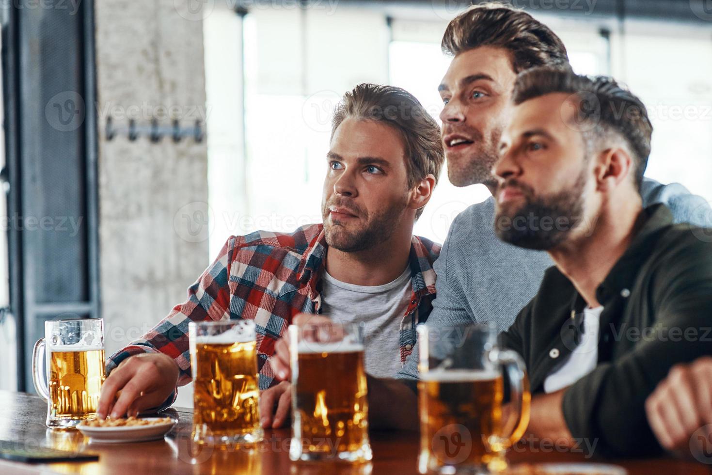 tre giovane uomini nel casuale capi di abbigliamento potabile birra e Guardando sport gioco mentre seduta nel il pub foto