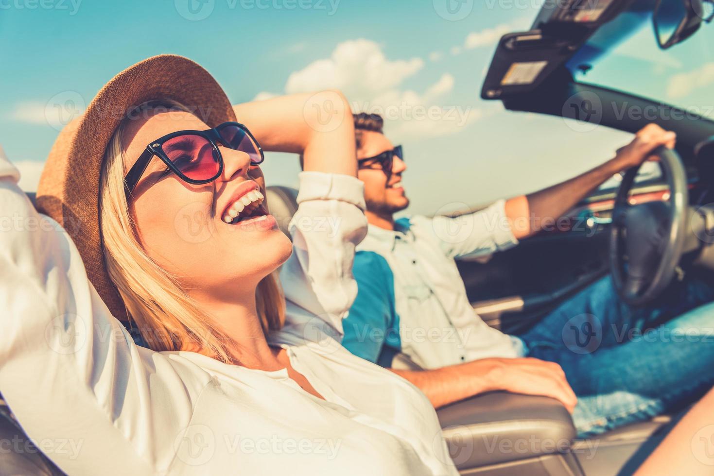 la libertà di il Aperto strada. lato Visualizza di gioioso giovane donna rilassante su il davanti posto a sedere mentre sua fidanzato seduta vicino e guida loro convertibile foto
