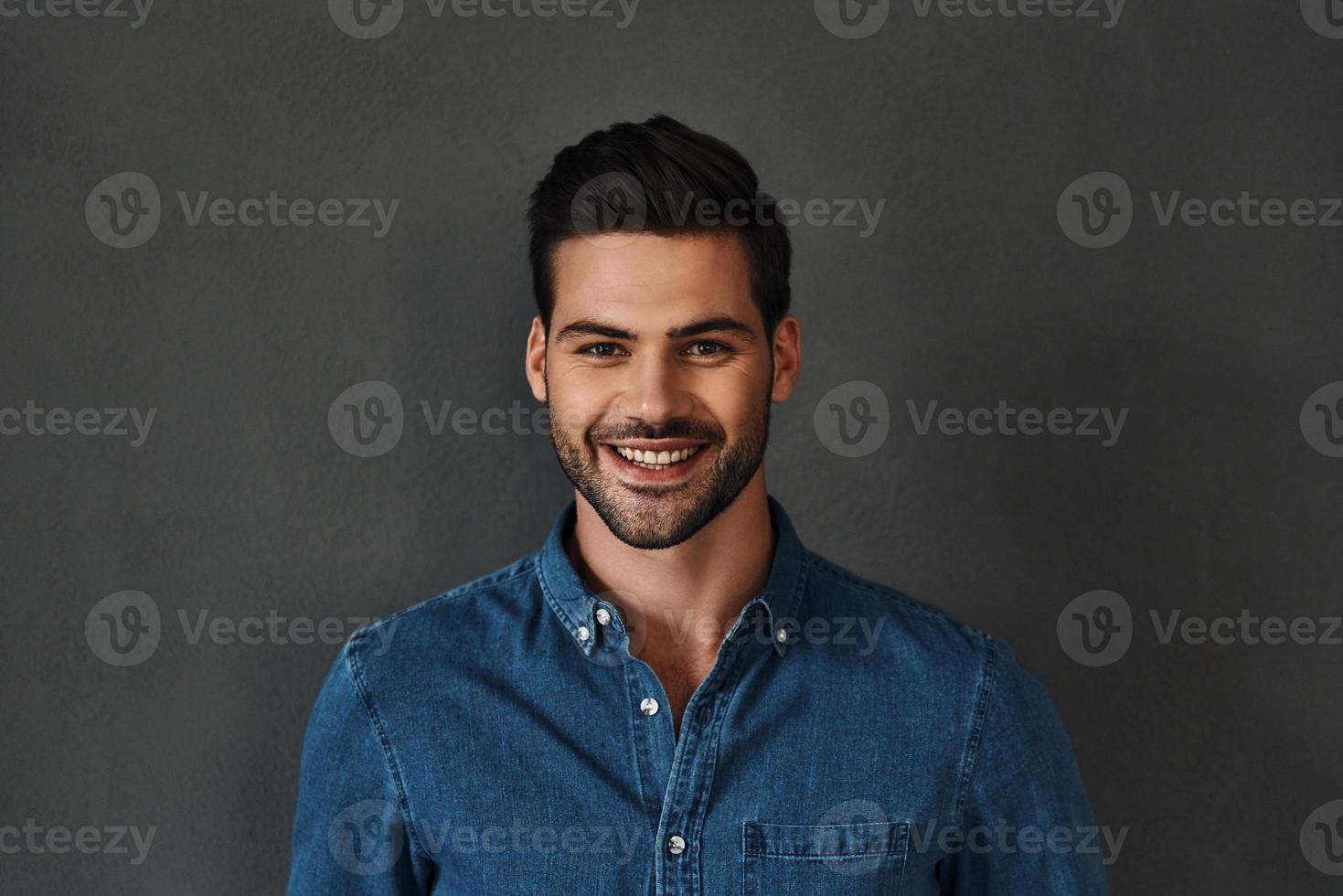 contento uomo. bello giovane uomo nel denim camicia guardare a telecamera e sorridente mentre in piedi contro grigio sfondo foto