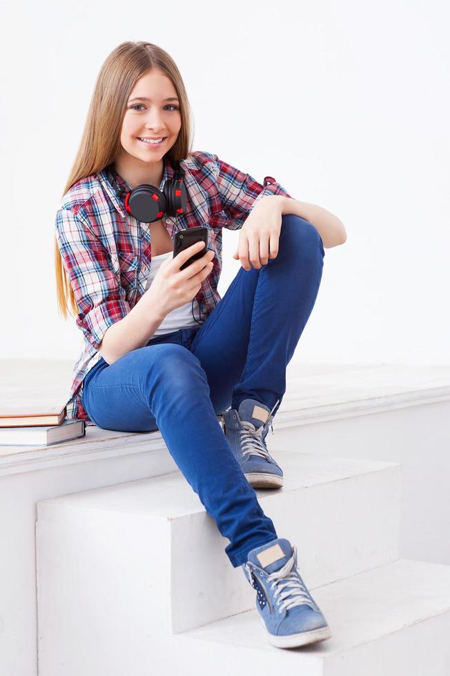 spensierato adolescente. carino adolescenziale ragazza Tenere mobile Telefono e sorridente mentre seduta su scala foto