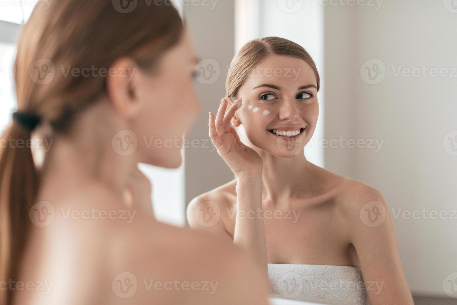 bellezza trattamento. al di sopra di il spalla Visualizza di giovane bellissimo giovane donna diffusione crema al di sopra di sua viso mentre guardare nel il specchio foto