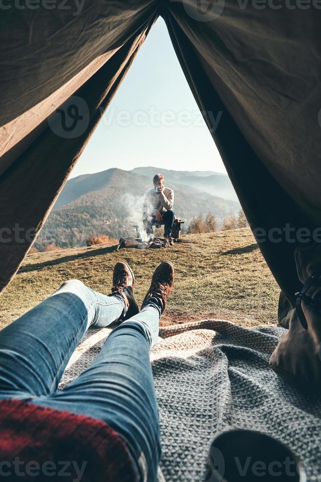 panoramico Visualizza. donna godendo il Visualizza di montagna gamma a partire dal il tenda mentre sua fidanzato riposo vicino il fuoco di bivacco foto