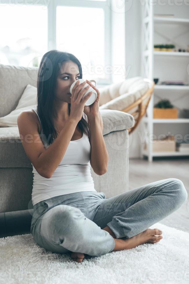 potabile caffè a casa. attraente giovane donna seduta su il tappeto a casa e potabile caffè foto