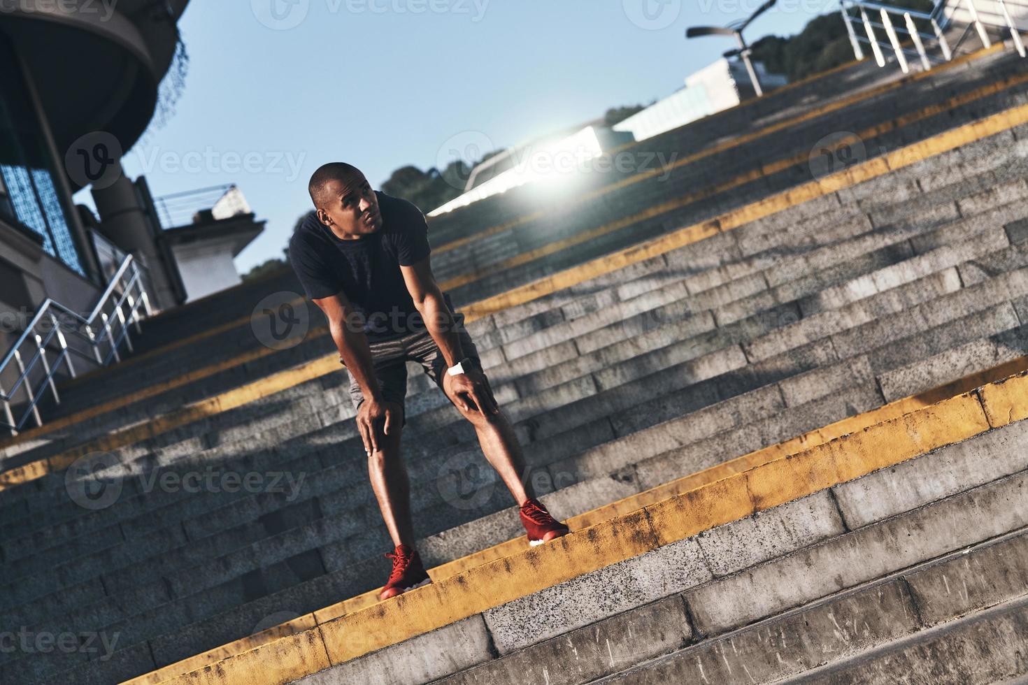 assunzione tempo per riposo. pieno lunghezza di giovane africano uomo nel gli sport capi di abbigliamento in piedi su il le scale mentre esercizio all'aperto foto
