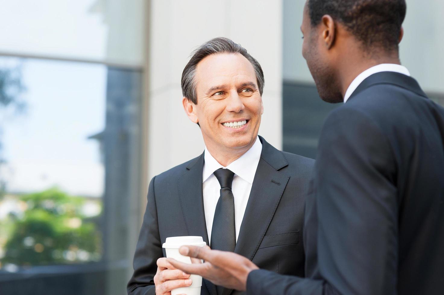 avendo un' caffè rompere per parlare intorno a. Due allegro attività commerciale uomini parlando e Esprimere a gesti mentre in piedi all'aperto foto