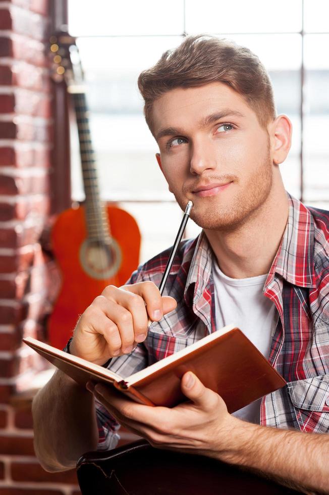 creativo dentro il suo anima. bello giovane uomo scrittura qualcosa nel Nota tampone e toccante il suo mento con penna mentre acustico chitarra posa nel il sfondo foto