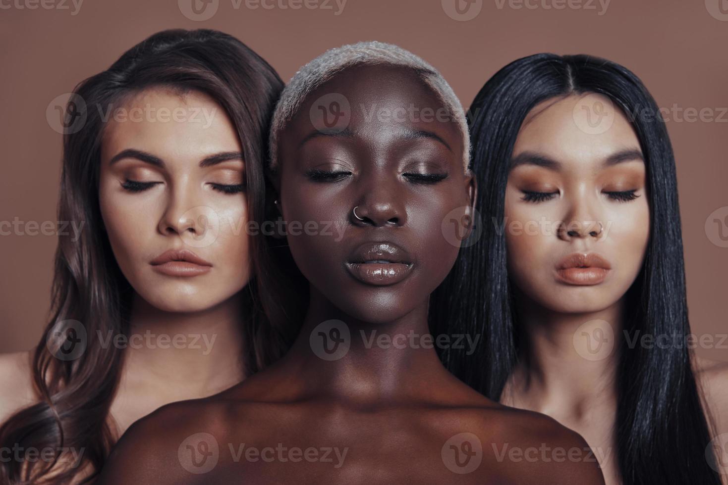 Perfetto pelle. tre attraente giovane donne conservazione occhi chiuso mentre in piedi contro Marrone sfondo foto