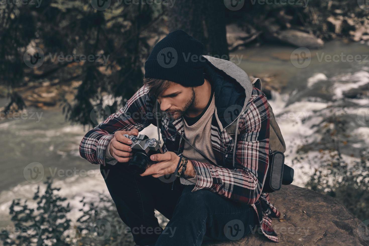 sempre trasporto telecamera. giovane moderno uomo con zaino Tenere un' foto telecamera mentre seduta nel il boschi con fiume nel sfondo