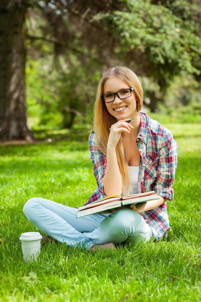 studiando su fresco aria. bellissimo giovane femmina alunno Tenere mano su mento e sorridente mentre seduta nel un' parco foto