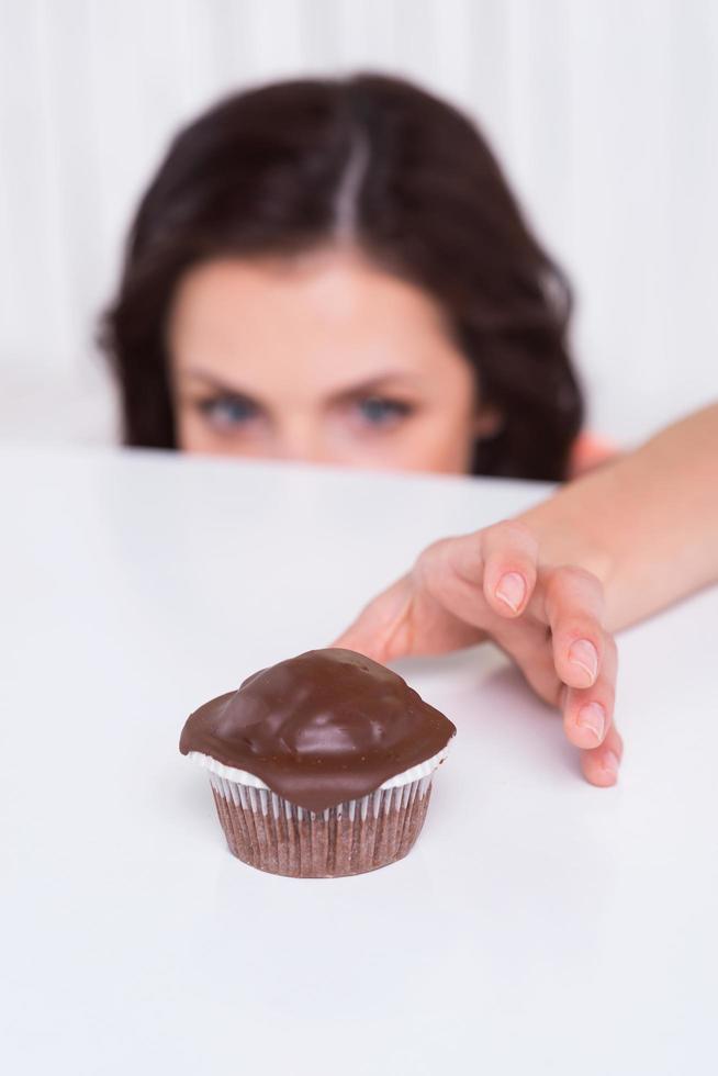 appena uno di più. giovane donna guardare su di il tavolo e allungamento mano per il cioccolato focaccina posa su il tavolo foto