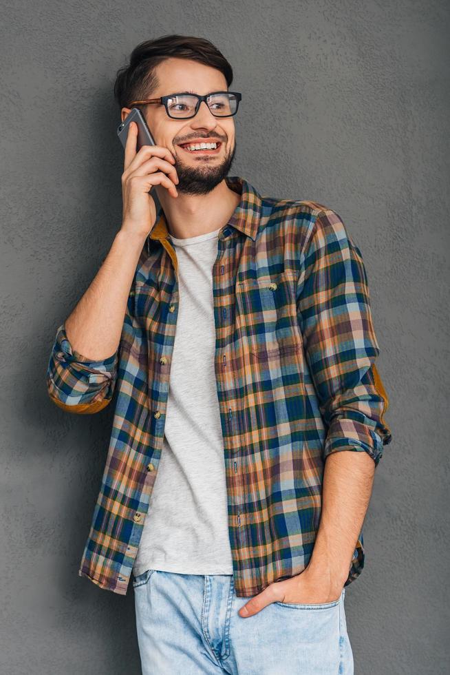 simpatico parlare. allegro giovane uomo nel bicchieri parlando su mobile Telefono e guardare lontano con Sorridi mentre in piedi contro grigio sfondo foto